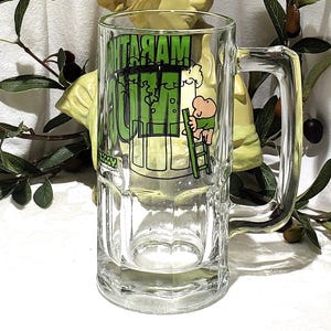 May include: Clear glass beer mug with a handle. The mug features a green and white graphic of a cartoon character climbing a ladder towards a beer tap, with the word "MARGARITA" in green.