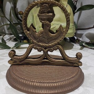 May include: A gold-toned metal anchor-shaped decorative stand with a rope-like design around the anchor. The stand has a round base with a brick-like pattern.