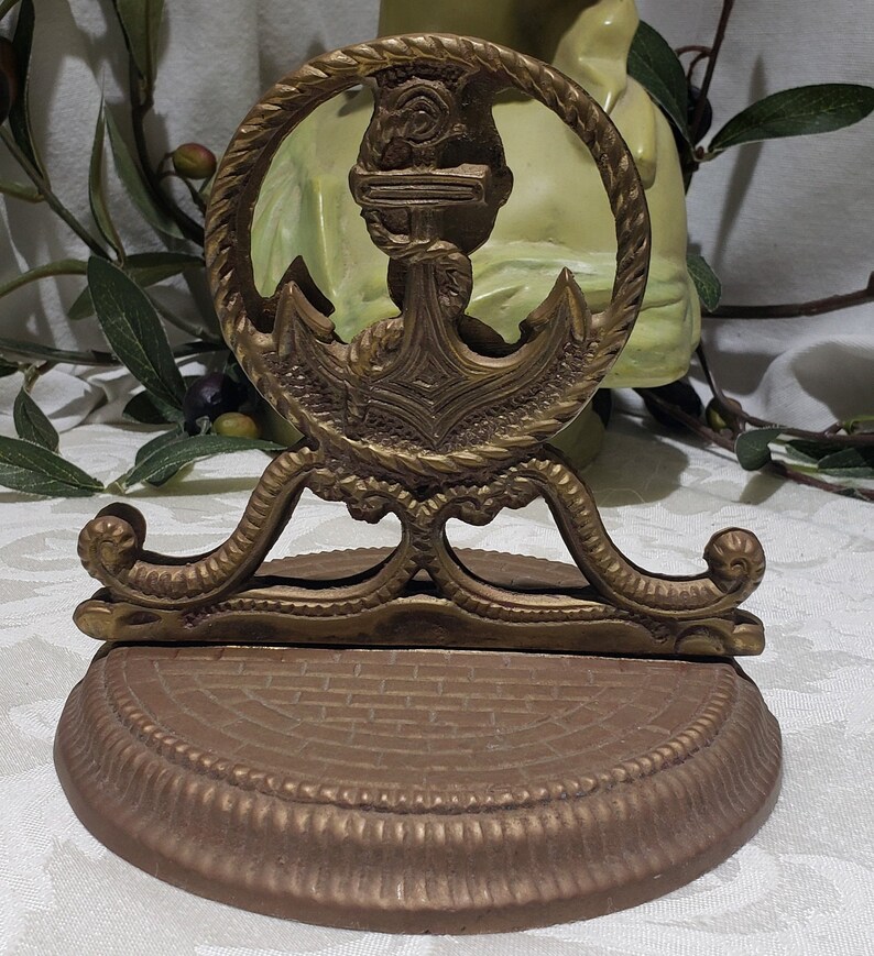 May include: A gold-toned metal anchor-shaped decorative stand with a rope-like design around the anchor. The stand has a round base with a brick-like pattern.
