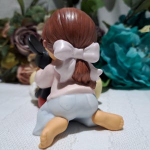 Pode incluir: Uma estatueta de cer&acirc;mica de uma crian&ccedil;a com cabelo castanho amarrado com um la&ccedil;o branco, vestindo uma camisa rosa e cal&ccedil;as azul claro, abra&ccedil;ando uma figura do Mickey Mouse. A estatueta est&aacute; em uma superf&iacute;cie branca.