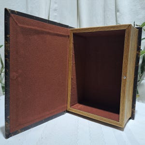 May include: An open book-shaped box with a dark brown exterior and a lighter brown interior. The box has a hinged lid and a small metal clasp. The interior is lined with a reddish-brown fabric.
