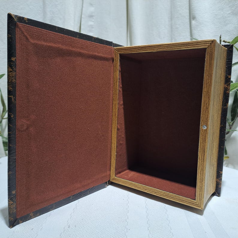 May include: An open book-shaped box with a dark brown exterior and a lighter brown interior. The box has a hinged lid and a small metal clasp. The interior is lined with a reddish-brown fabric.
