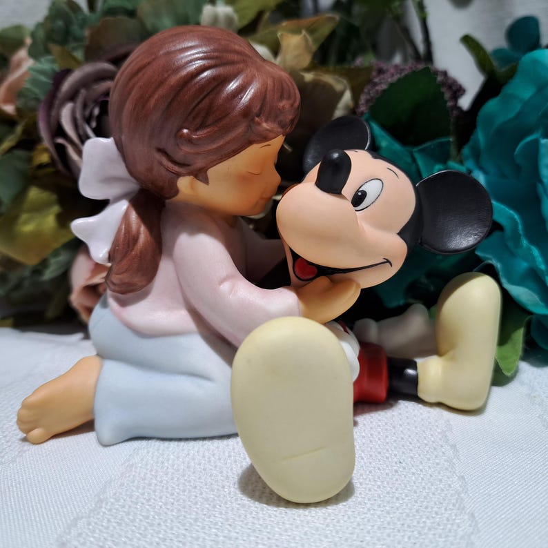 Pode incluir: Uma estatueta de cer&acirc;mica de uma menina com uma camisa rosa e saia azul, beijando uma cabe&ccedil;a do Mickey Mouse. A estatueta tem um la&ccedil;o branco no cabelo e est&aacute; sentada em uma superf&iacute;cie branca. Mickey Mouse tem orelhas pretas e boca vermelha.
