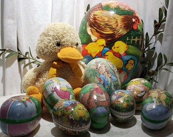 May include: A collection of vintage Easter egg-shaped tins with various colorful illustrations. The tins feature a hen with chicks, a rabbit, and other whimsical designs. A plush yellow duck sits next to the tins.