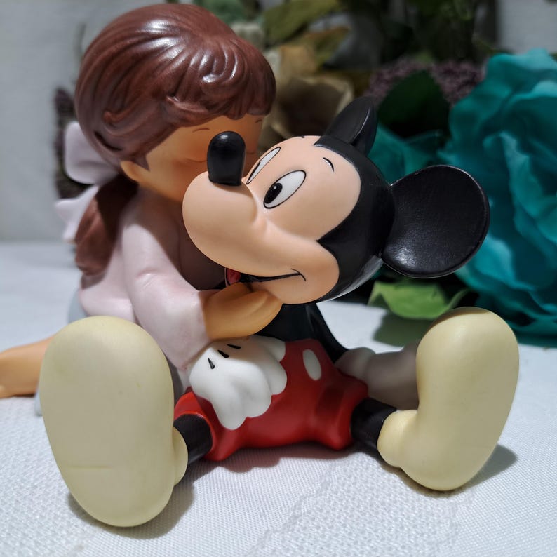 Pode incluir: Uma estatueta de uma menina com um vestido rosa abra&ccedil;ando uma figura do Mickey Mouse. A figura do Mickey Mouse est&aacute; usando shorts vermelhos, sapatos pretos e luvas brancas. A menina tem cabelo castanho e um la&ccedil;o branco.