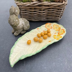 May include: A ceramic leaf-shaped serving dish with a floral design and yellow sweets. A small grey rabbit figurine and a wicker basket filled with green plants are in the background.