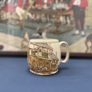 May include: A white ceramic mug with a brown and white illustration of a building with the text "George Inn" on a sign. The mug has a handle on the right side.