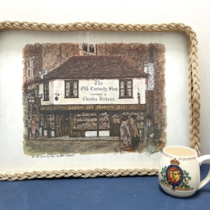 May include: A woven wicker tray featuring a printed image of a shop called "The Old Curiosity Shop", made famous by Charles Dickens. The shop is depicted in a vintage style with antique and modern art displayed in the windows. The tray is on a blue surface next to a white ceramic mug with a royal crest.