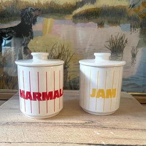 May include: Two white ceramic jars with lids. One jar has the word "MARMAL" in red, the other has "JAM" in yellow. Both jars have vertical stripes. The background features a framed painting of a dog and a landscape.