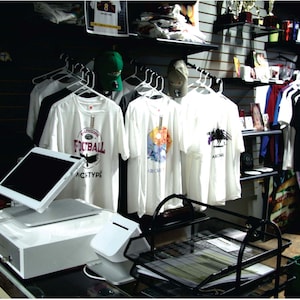 May include: A retail store interior with white t-shirts hanging on display. The shirts have various graphic designs and text. A point-of-sale system with a tablet is on the counter. A lava lamp is visible on the left.