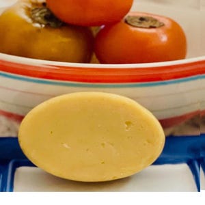 May include: A smooth, oval-shaped pale yellow bar of soap positioned in the foreground. Behind the soap sits a white bowl with horizontal red and blue stripes, containing three round, orange and yellow persimmon fruits with brown stems.