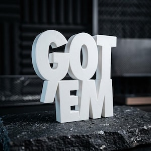 May include: A white, three-dimensional sign featuring the text GOT EM in a bold, block-letter font. The letters are arranged in two rows, with GOT on the top and EM on the bottom, creating a sturdy, freestanding display piece with a matte finish.
