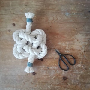 A button knot made by the elephant of joy from white cotton rope, with a greeny-grey cord on the loose ends. The knot sits on a distressed wooden background with a pair of black utility scissors next to it