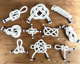 Könnte beinhalten: Zehn verschiedene Knotenarten, die mit weißem Seil gebunden sind, jeweils mit blauen Akzenten an den Enden. Die Knoten werden auf einer Holzoberfläche gezeigt.