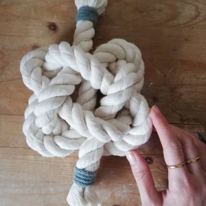 A button knot made by the elephant of joy from white cotton rope, with a greeny-grey cord on the loose ends. The knot sits on a distressed wooden background with a white female hand holding it