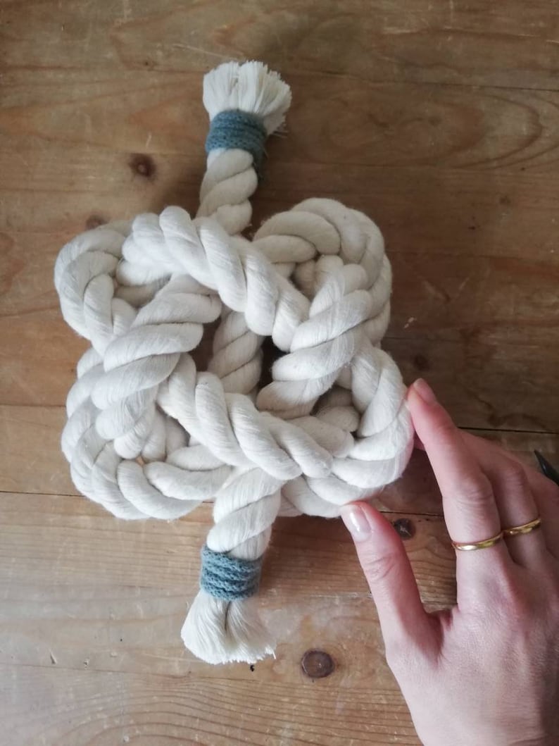A button knot made by the elephant of joy from white cotton rope, with a greeny-grey cord on the loose ends. The knot sits on a distressed wooden background with a white female hand holding it