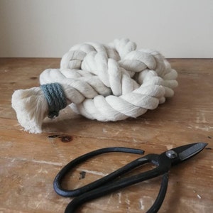 A low angled photo of button knot made by the elephant of joy from white cotton rope, with a greeny-grey cord on the loose ends. The knot sits on a distressed wooden surface against a cream wall with a pair of black utility scissors next to it