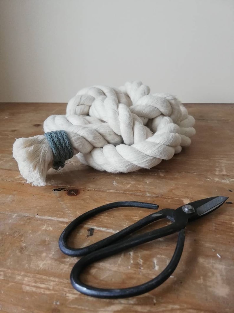 A low angled photo of button knot made by the elephant of joy from white cotton rope, with a greeny-grey cord on the loose ends. The knot sits on a distressed wooden surface against a cream wall with a pair of black utility scissors next to it