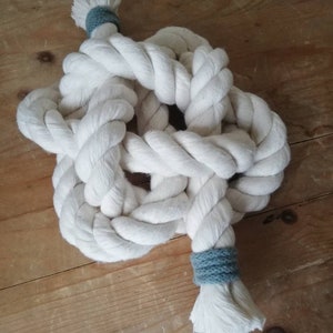 An angled image of a button knot made by the elephant of joy from white cotton rope, with a greeny-grey cord on the loose ends. The knot sits on a distressed wooden background with a pair of black utility scissors next to it