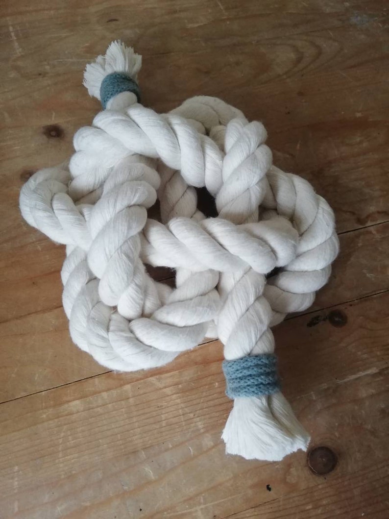 An angled image of a button knot made by the elephant of joy from white cotton rope, with a greeny-grey cord on the loose ends. The knot sits on a distressed wooden background with a pair of black utility scissors next to it