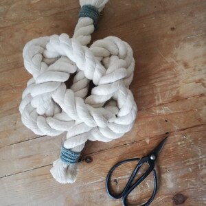 A button knot made by the elephant of joy from white cotton rope, with a greeny-grey cord on the loose ends. The knot sits on a distressed wooden background with a pair of black utility scissors next to it