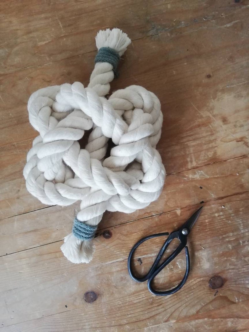 A button knot made by the elephant of joy from white cotton rope, with a greeny-grey cord on the loose ends. The knot sits on a distressed wooden background with a pair of black utility scissors next to it