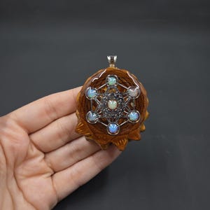 May include: A circular pendant featuring a brown, textured pinecone base with a silver-toned metal geometric Metatron's Cube overlay. Seven small, iridescent, round opals are set at the intersections of the geometric lines, with a silver bail.