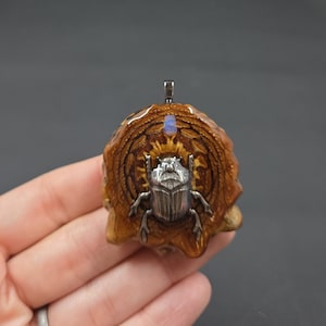 May include: A handcrafted pendant featuring a silver scarab beetle on a carved wooden base. The pendant has a small, iridescent blue stone at the top. The wooden base is a warm brown color, with intricate carved details.