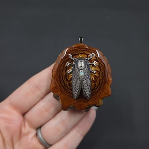 May include: A handcrafted pendant featuring a detailed silver cicada charm with an iridescent stone, set on a carved, brown wooden base. The pendant has a small loop for a chain. The design showcases natural textures and insect-inspired artistry.