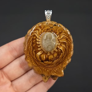 May include: A pendant featuring a fossilized coral encased in resin, with a detailed, intricate design around the coral. The pendant is a light brown color with a silver bail.