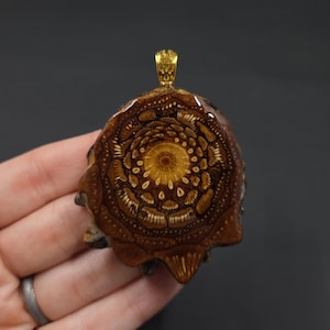 May include: A handcrafted pendant with a unique design. The pendant features a detailed, circular pattern in shades of brown and gold, with a gold-colored bail at the top. The pendant is made of wood and resin, showcasing natural textures and colors.