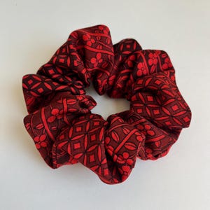 Puede incluir: Una goma para el pelo burdeos con un patrón geométrico y floral repetido. La tela es de color rojo oscuro con un diseño rojo más oscuro. La goma para el pelo está fruncida y tiene un aspecto suave y texturizado. Este accesorio para el cabello es adecuado para varios peinados.