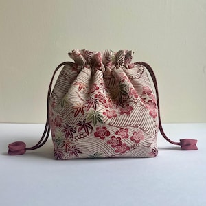 May include: A drawstring pouch featuring a cream-colored fabric with a repeating pattern of red and green maple leaves and plum blossoms, accented by wavy lines. The bag is cinched at the top with two dark red braided cords with knotted ends.