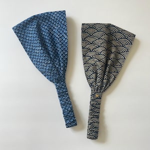 Może przedstawiać: Dwie materiałowe opaski do włosów. Jedna jest granatowa z białym wzorem w kropki. Druga jest czarna z beżowym wzorem fal. Obie opaski mają szeroki, marszczony design i wąski, zapinany pasek.