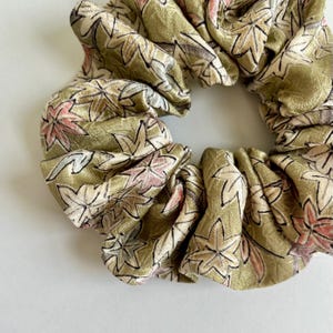 May include: A close-up of a scrunchie with a floral pattern. The scrunchie is a light olive green color with a repeating pattern of leaves in shades of pink, beige, and black. The fabric has a slight sheen.