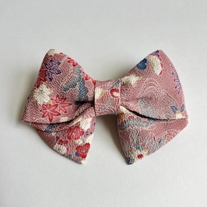 May include: A bow-shaped hair accessory made of textured fabric in a dusty rose color. The material features a repeating pattern of red, white, purple, and blue floral and leaf motifs, along with subtle gray wave-like line details throughout.