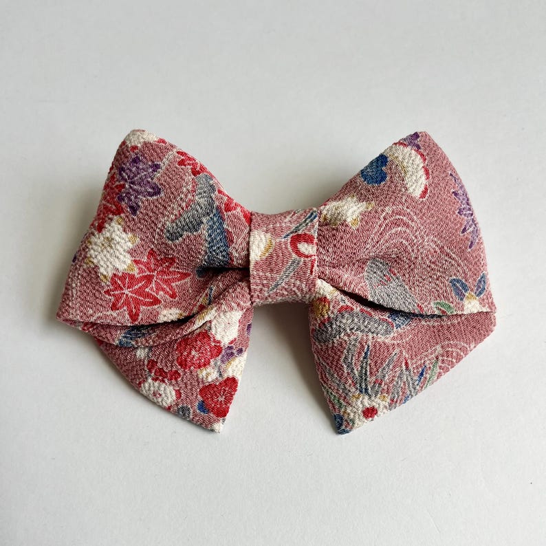 May include: A bow-shaped hair accessory made of textured fabric in a dusty rose color. The material features a repeating pattern of red, white, purple, and blue floral and leaf motifs, along with subtle gray wave-like line details throughout.