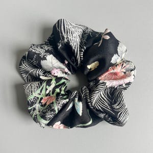 May include: A black fabric hair scrunchie featuring a botanical print with white fern leaves, pink flowers, and green stems. The accessory has a gathered, ruffled texture and a smooth, slightly reflective finish throughout the circular design.