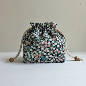 Könnte beinhalten: Ein kleiner Beutel mit Kordelzug und floralem Muster. Der Stoff ist marineblau mit einem sich wiederholenden Muster aus kleinen weißen, rosa und grünen Blumen. Der Kordelzug ist ein beigefarbenes Seil mit einer Holzperle an jedem Ende.