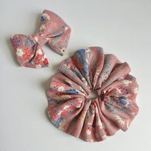 May include: A dusty pink fabric hair accessory set consisting of a bow hair clip and a circular scrunchie. Both items feature a textured surface with a repeating pattern of small white, red, blue, and purple floral and wave-like graphic motifs.