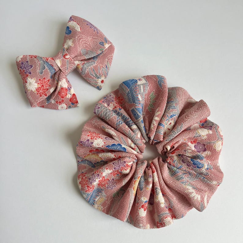May include: A dusty pink fabric hair accessory set consisting of a bow hair clip and a circular scrunchie. Both items feature a textured surface with a repeating pattern of small white, red, blue, and purple floral and wave-like graphic motifs.