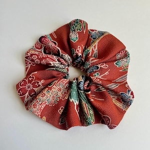 May include: A fabric hair scrunchie in a deep rust-red color featuring a repeating floral pattern in shades of white, green, and blue. The material has a textured, crepe-like surface with gathered folds creating a circular, voluminous shape.