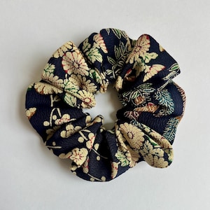 May include: A dark navy blue fabric hair scrunchie featuring a repeating pattern of cream-colored chrysanthemum flowers with subtle green and red accents. The material has a textured, crepe-like surface with gathered folds throughout the circular design.