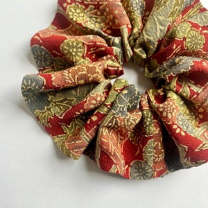 May include: A scrunchie made of fabric with a floral pattern in shades of red, green, and gold. The scrunchie is gathered and has a circular shape, suitable for various hairstyles.