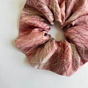 May include: A pink and white patterned scrunchie made of silky fabric. The scrunchie has a white geometric pattern on a pink background.