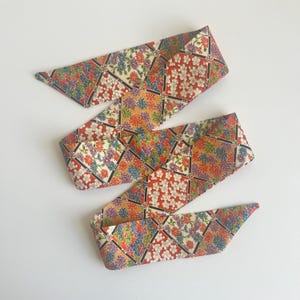 May include: A long, narrow fabric ribbon featuring a repeating diamond pattern filled with colorful floral motifs. The design includes clusters of red, blue, yellow, and white flowers against a cream background, separated by thin black diagonal lines.
