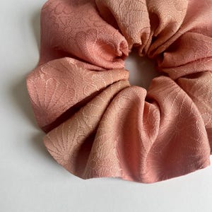 May include: Close-up of a large, peach-colored scrunchie. The fabric has a subtle floral pattern. The scrunchie is gathered and folded, creating a voluminous, textured appearance. The background is plain white.