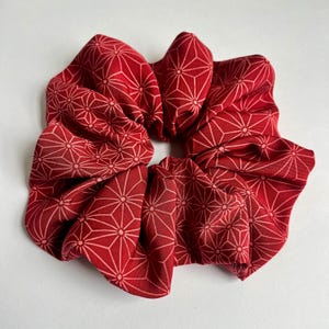 May include: A fabric hair scrunchie in a deep red colour featuring a repeating geometric pattern of light pink lines forming interconnected star-like shapes. The accessory has a gathered, ruffled texture with a circular opening in the centre.