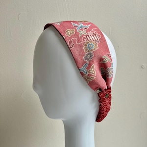 May include: A red fabric headband with a floral and bird pattern. The headband is tied at the base and features a smaller, patterned section. The design includes blue, yellow, and white accents.