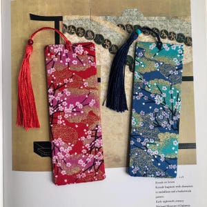 以下が含まれることがあります： タッセル付きの布製ブックマーク2つ。左側のブックマークは、ピンクとゴールドのアクセントが施された赤色で、赤いタッセルが付いています。右側のブックマークは、緑とゴールドのアクセントが施された青色で、青いタッセルが付いています。両方のブックマークは、白い花とゴールドのアクセントが施された花柄です。